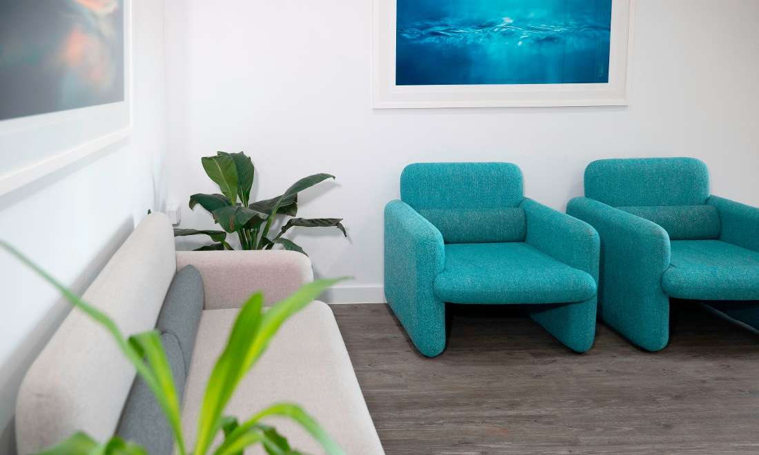 The waiting room at Waterways Dental. A grey lounge and two blue arm chairs along two white walls. Imagery of water on both walls. 